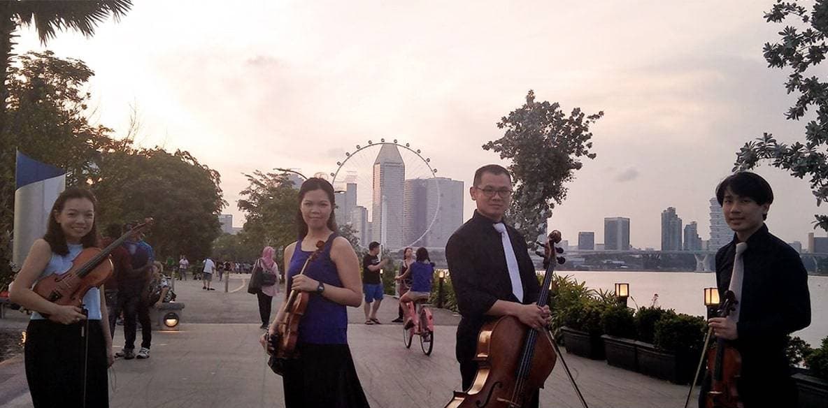 The GraceNotes string quartet standing by the water at Marina Bay at dusk, with the Singapore skyline behind them